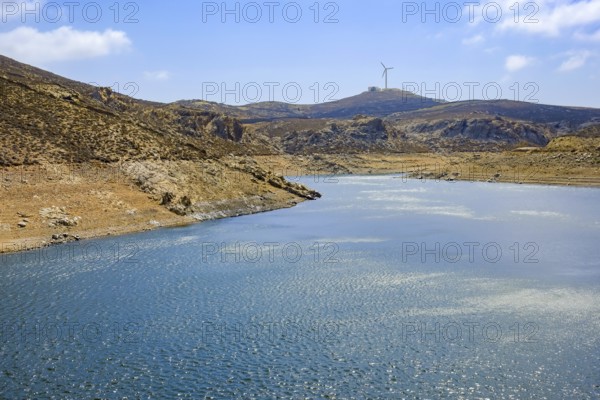 Mykonos, Cyclades, Greece - Dry Fokos Reservoir near Ano Mera. The Mykonos Municipal Water Supply and Sewage Management Company has two dams, Marathi Reservoir and Fokos Reservoir, to supply water to the island. Due to the prolonged drought, both are almost empty and are not being used. A wind turbine at the back of the mountain