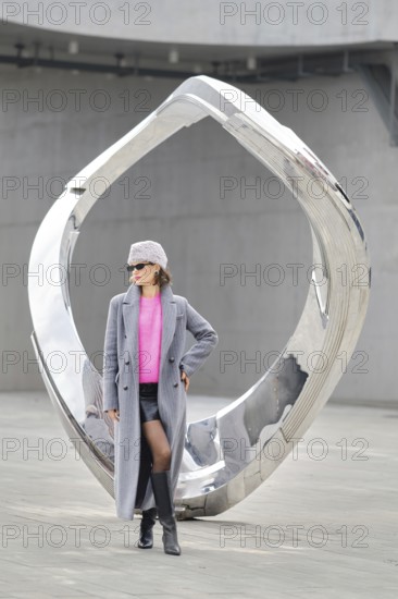 A woman stands confidently in urban attire against a shiny modern sculpture. The scene showcases vibrant street fashion with a trendy pink top, gray coat, and stylish boots