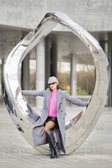 A stylish woman poses sitting on metallic sculpture in a city, showcasing urban street fashion