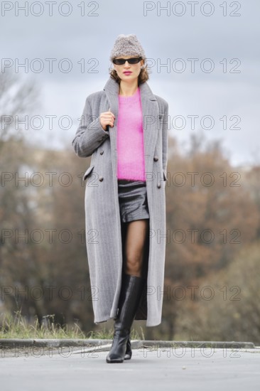 A stylish woman walks down a city street, wearing a pink sweater, unbuttoned long coat, and leather boots. The background features trees and cloudy skies, creating a dynamic urban atmosphere