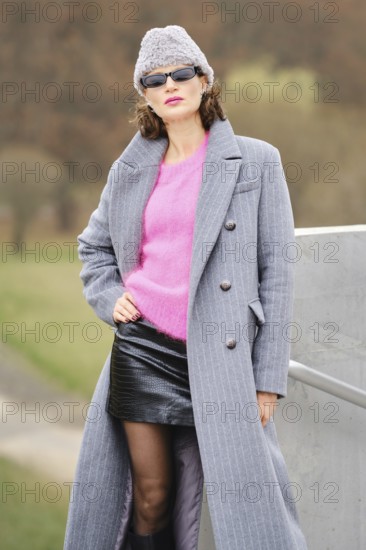 A woman stands playfully in city park in a stylish autumn outfit, showcasing urban street style. She wears a pink sweater and a sleek black skirt, paired with trendy sunglasses and a cozy beanie