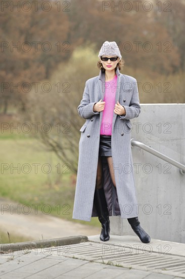 A woman showcases striking urban fashion by wearing a bright pink sweater, a long gray coat, and sleek black boots. She stands confidently on a concrete structure at a park during the daytime