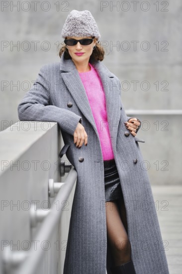 A woman stands confidently on an urban street, dressed in a long gray coat over a vibrant pink sweater. She pairs her outfit with stylish sunglasses and a cozy beanie hat, showcasing modern fashion