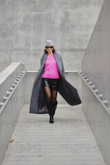 A fashionable woman in sunglasses rushing down a sleek grey walkway, wearing a mini leather skirt, boots, pink sweater and a hat. Her unbuttoned long coat flutters in the wind