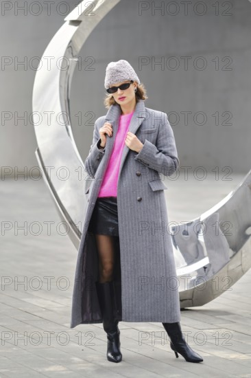 A woman in a long gray coat and pink sweater posing near a large modern sculpture. She wears black boots and hat, showcasing urban fashion in city setting