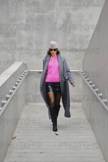 A stylish woman walks down a sleek urban walkway, dressed in a stylish gray coat over a bright pink sweater and black leather skirt. The scene captures a modern aesthetic under natural light