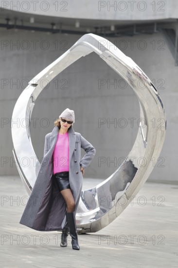 A woman wearing a vibrant pink sweater and gray coat stands next to a shiny modern sculpture. The urban setting captures the essence of street style during daylight hours
