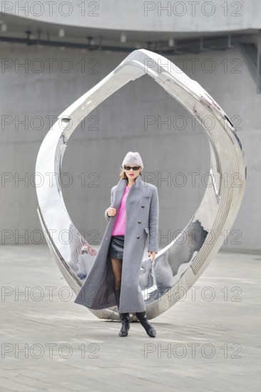 A stylish woman wears a grey coat and pink sweater while posing by a sleek, reflective sculpture in an urban setting