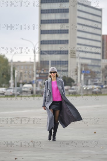 A stylish woman walks along a city sidewalk, wearing a vibrant pink sweater, leather skirt and a grey coat. The urban backdrop features modern buildings and bustling traffic, creating a lively scene