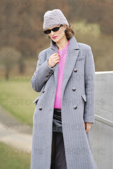 A stylish woman in a long gray coat and bright pink sweater stands outdoors. She wear sunglasses and triangular fur beanie hat while posing against autumn trees and grass