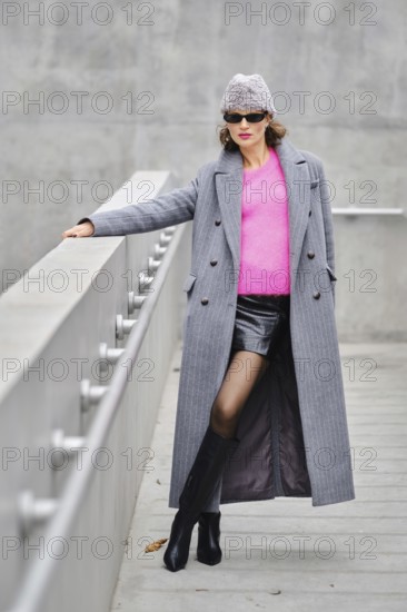 A stylish playful woman poses on a contemporary walkway, wearing a long gray coat, pink sweater, and black mini skirt