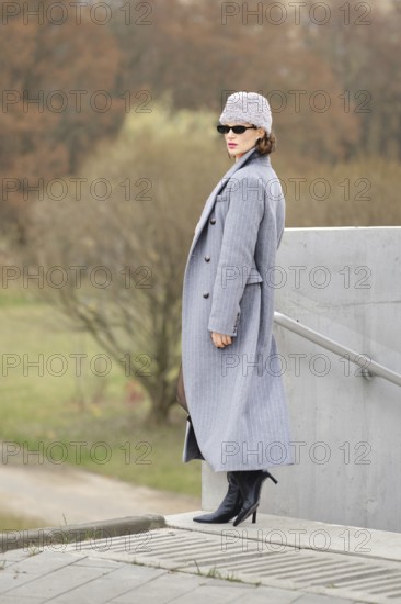 A fashionable woman poses on steps wearing a long grey coat, high-heeled boots, and a stylish beanie hat. The background features a blurred park with trees and a pathway, adding an urban flair