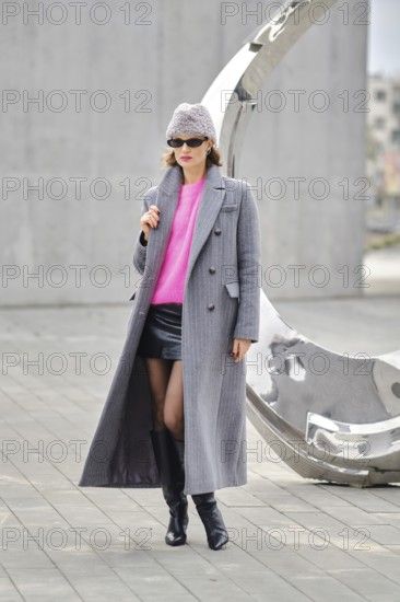 In an urban setting, a woman posing in a long gray coat over a pink sweater and a black leather mini skirt. She complemented her look with a triangular fur hat, sunglasses and knee-high boots