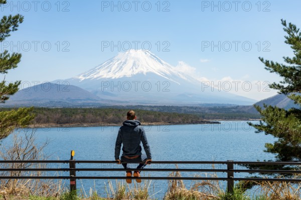 Young man sitting on railings next to a road and looking across the lake to Mt Fuji volcano, Motosu Lake, Yamanashi Prefecture, Japan