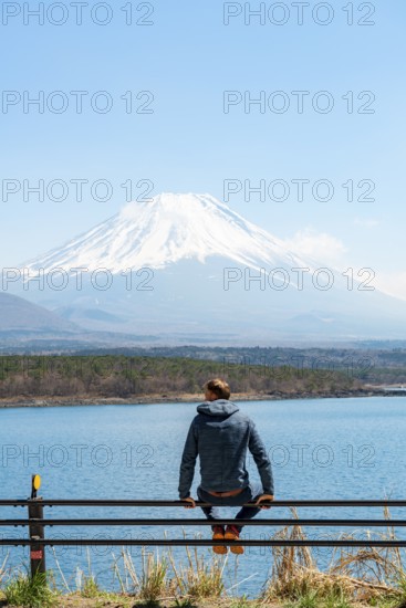 Young man sitting on railings next to a road and looking across the lake to Mt Fuji volcano, Motosu Lake, Yamanashi Prefecture, Japan