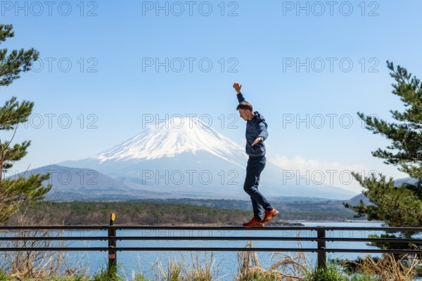 Young man balancing on railings next to a road, view across the lake to Mt Fuji volcano, Motosu Lake, Yamanashi Prefecture, Japan