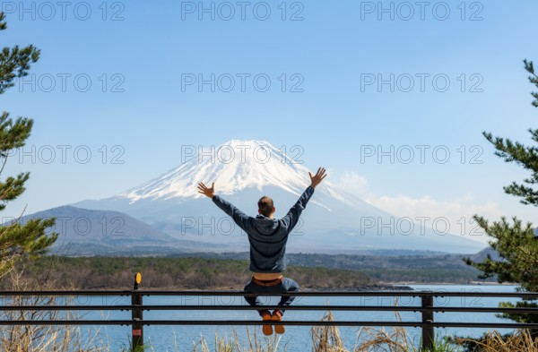 Young man sitting on railings next to a road and stretching his arms in the air, looking across the lake to Mt Fuji volcano, Motosu Lake, Yamanashi Prefecture, Japan