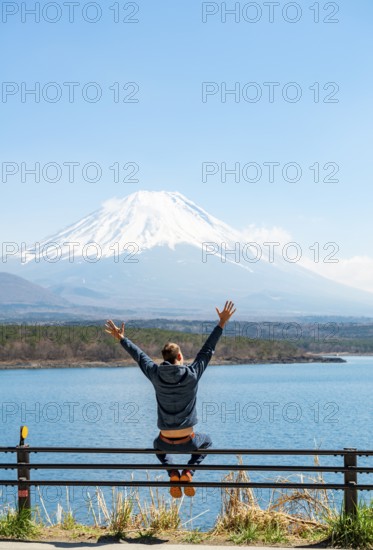 Young man sitting on railings next to a road and stretching his arms in the air, looking across the lake to Mt Fuji volcano, Motosu Lake, Yamanashi Prefecture, Japan