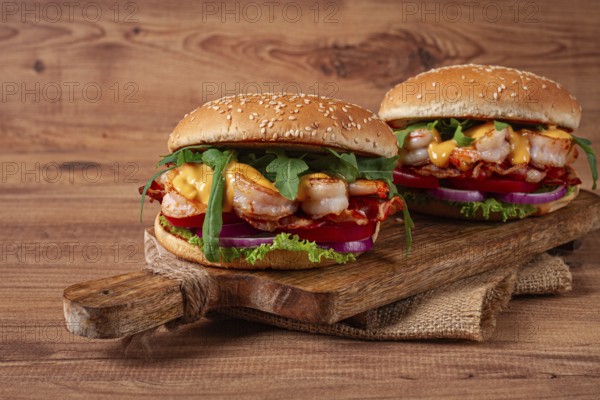 Two shrimp burgers and buns with sesame seeds, bacon, lettuce, tomatoes and onions, on a wooden board, rustic style