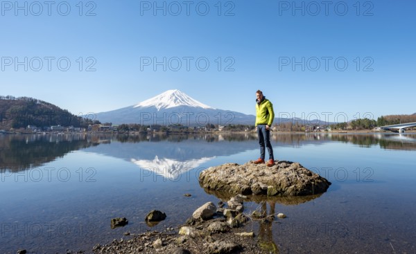 Young man standing on a rock in water, volcano Mt. Fuji is reflected in Lake Kawaguchi, Yamanashi Prefecture, Japan