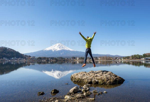 Young man standing on a rock in water, jumping into the air, volcano Mt. Fuji is reflected in Lake Kawaguchi, Yamanashi Prefecture, Japan