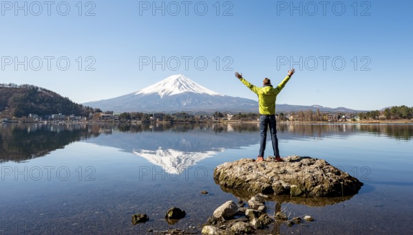 Young man stands on a rock in water, stretches his arms in the air, volcano Mt. Fuji is reflected in Lake Kawaguchi, Yamanashi Prefecture, Japan