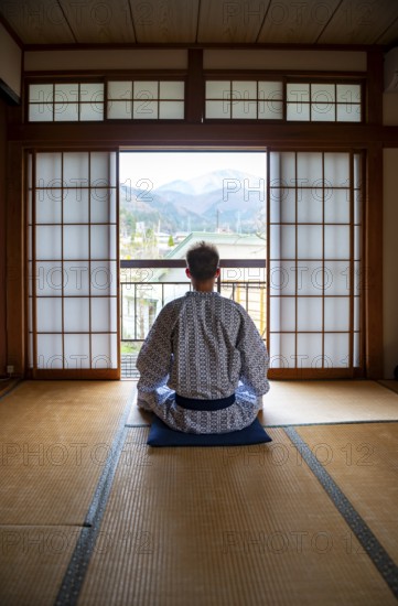 Young man wearing kimono sitting in traditional Japanese living room with tatami mats and shoji sliding doors, from behind, Yamanouchi, Nagano, Japan