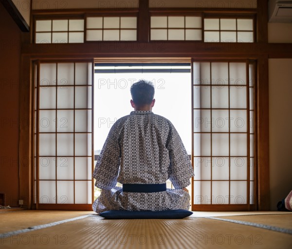 Young man wearing kimono sitting in traditional Japanese living room with tatami mats and shoji sliding doors, from behind, Yamanouchi, Nagano, Japan