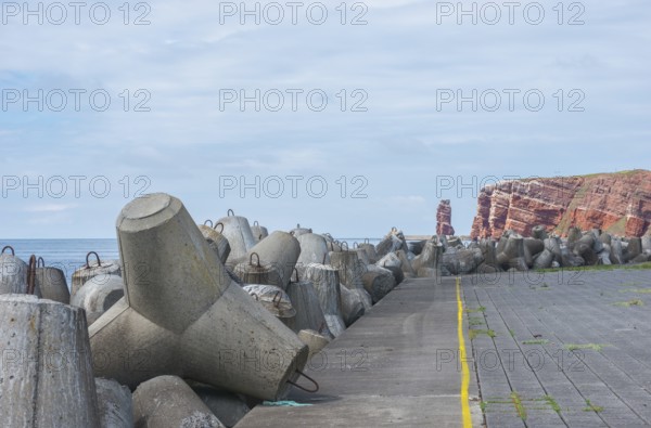 Coastline with concrete tetrapods at sea, coastal protection, protective wall, paved coastal strip with yellow line, danger, marking, warning, red cliffs, cliff, cliffs, rock layers in the red sandstone and Lange Anna in the background, surf pillar, escape line, perspective, overcast sky, blue sea on the horizon, high-seas island of Heligoland, North Sea, Pinnland district Berg, Schleswig-Holstein, Germany
