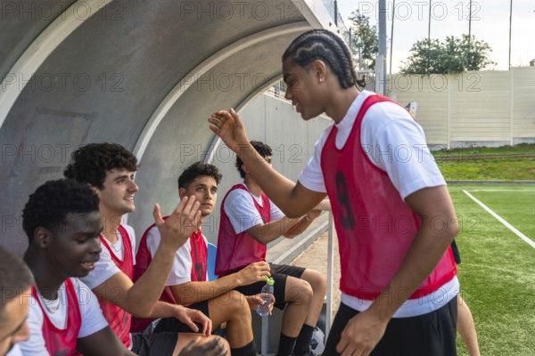 Soccer players in red bibs sitting on the bench and high fiving in the dugout, sharing a moment of camaraderie and teamwork during a game on the football field