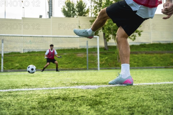 Soccer player's leg striking a football during a penalty kick on a green artificial turf field, with a goal and a waiting goalkeeper in the background