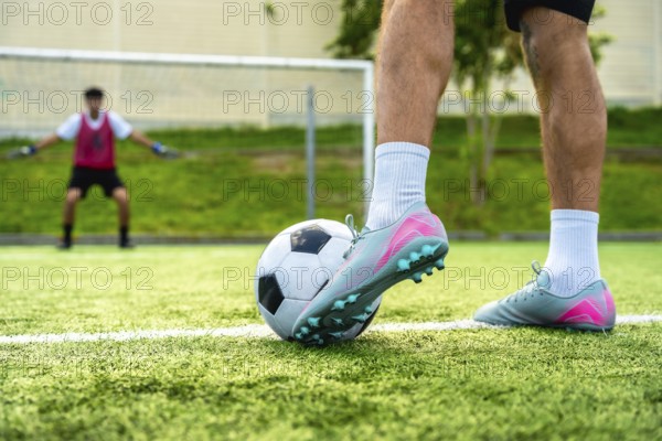 Soccer player's foot rests on a football, ready to take a penalty shot on a green artificial turf field, with a blurred goalkeeper standing in the background blocking the goal