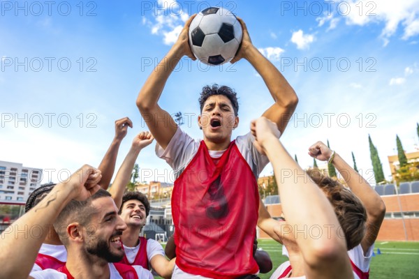 Young men soccer teammates in red bibs celebrate victory, raising a ball overhead with joyful cheers on a sunny stadium field, capturing teamwork and triumphant energy