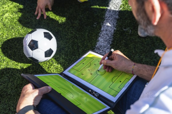 Soccer coach sketching tactical game plan on a clipboard beside a ball on artificial grass near a white sideline, focused on training, teamwork and match strategy