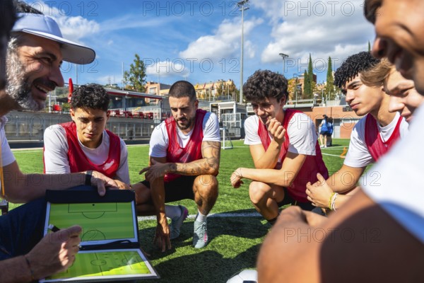 Soccer coach outlining a game strategy on a clipboard with a marker while adult and young male players in red bibs listen attentively during training on a green artificial turf field