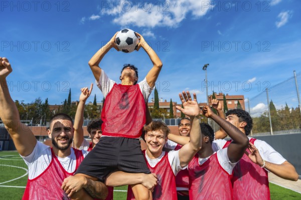 Young male soccer team members celebrating a goal or game victory on a green athletic field, lifting one player holding the ball high under a blue sky, all wearing red bibs