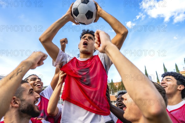 Young diverse men's soccer team members are cheering loud with fists raised and one player is triumphantly lifting a football overhead, symbolizing unity and achievement