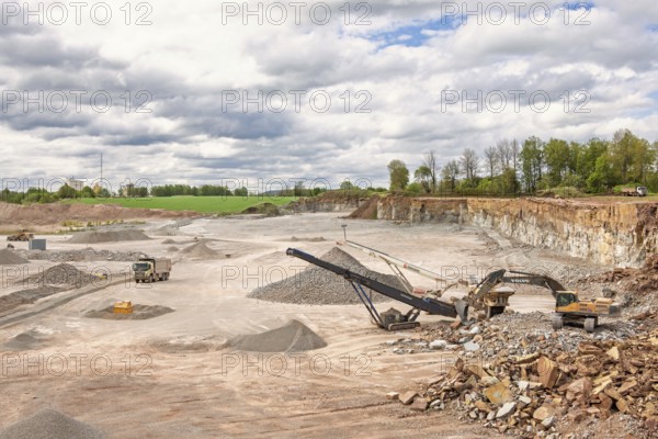 Excavator at a stone crusher with a conveyor in a limestone quarry with a waiting truck in a rural landscape, Sweden