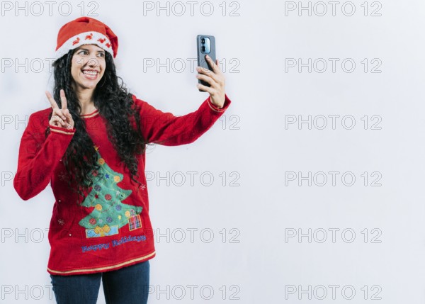 Happy young latin woman in Christmas sweater taking selfies isolated. Funny girl in Christmas hat and sweater taking selfie, isolated