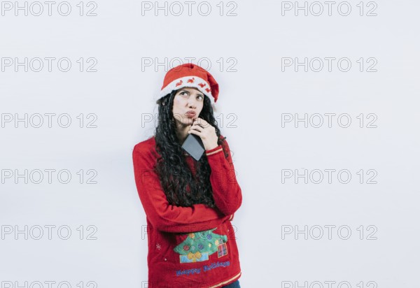 Young woman in christmas sweater thoughtful holding cell phone isolated. Girl in christmas hat thinking holding phone