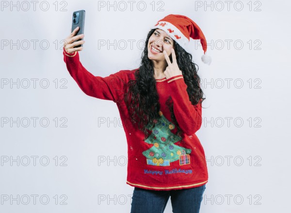 Funny girl in Christmas hat and sweater taking selfie, isolated. Happy young latin woman in Christmas sweater taking selfies isolated