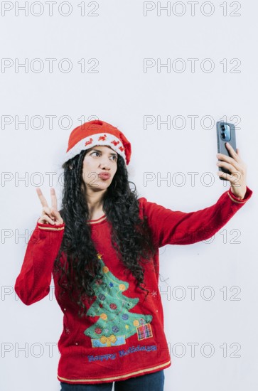Funny latin girl in Christmas hat taking selfies and making gestures isolated. Cheerful young woman in Christmas sweater taking selfies