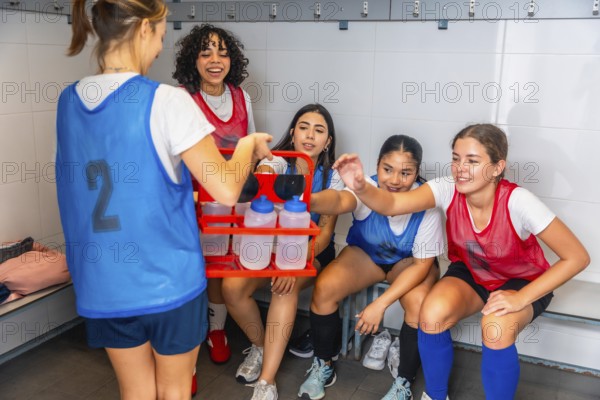 Teenagers from a diverse girls soccer team taking a water break and hydrating, showing friendship, sports, and teamwork in the locker room after practice or a game