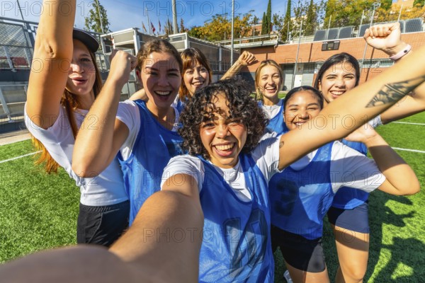 Diverse group of young women soccer players celebrating an achievement or victory, taking a joyful selfie together on a green artificial turf field, showing teamwork and friendship