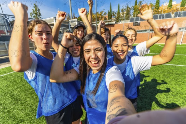 Happy young women's soccer team members celebrate a sports victory together on the green turf, raising their fists in excitement and taking a happy group selfie