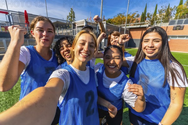 Group of smiling young women players celebrating success and victory on a soccer field, posing for a selfie after a match, showcasing teamwork and friendship