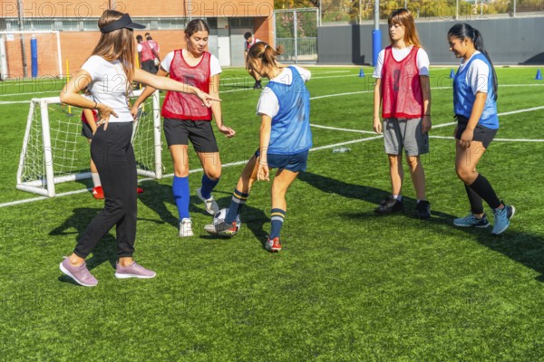 Female coach instructing a diverse group of young women soccer players on a green artificial turf field, practicing football skills and working on teamwork during sports training