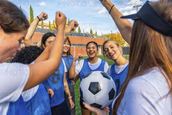Happy female soccer team players huddling together on the field, raising their fists in celebration, and holding a football, representing teamwork and success
