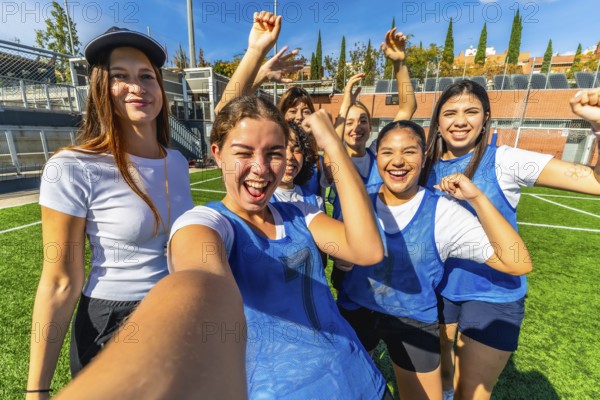 Group of young, multiracial women soccer players cheering and taking a joyful selfie on sunny green turf after a victory, arms raised in celebration and friendship