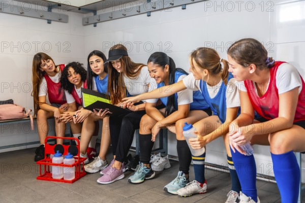 Diverse women's soccer team in uniforms sitting on benches in a locker room, gathered around their coach studying a clipboard as they plan strategy and prepare for the game